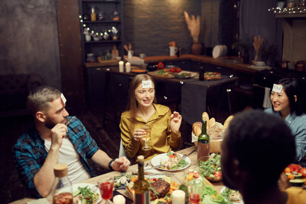 Multi-ethnic group of young people playing guessing game while sitting at table during dinner party in dark room, copy space